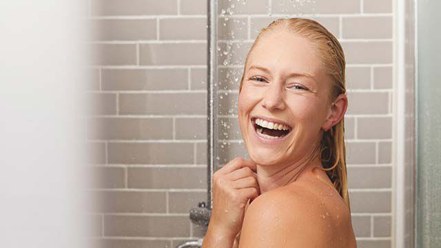 Blonde Frau steht lachend unter der Dusche 
